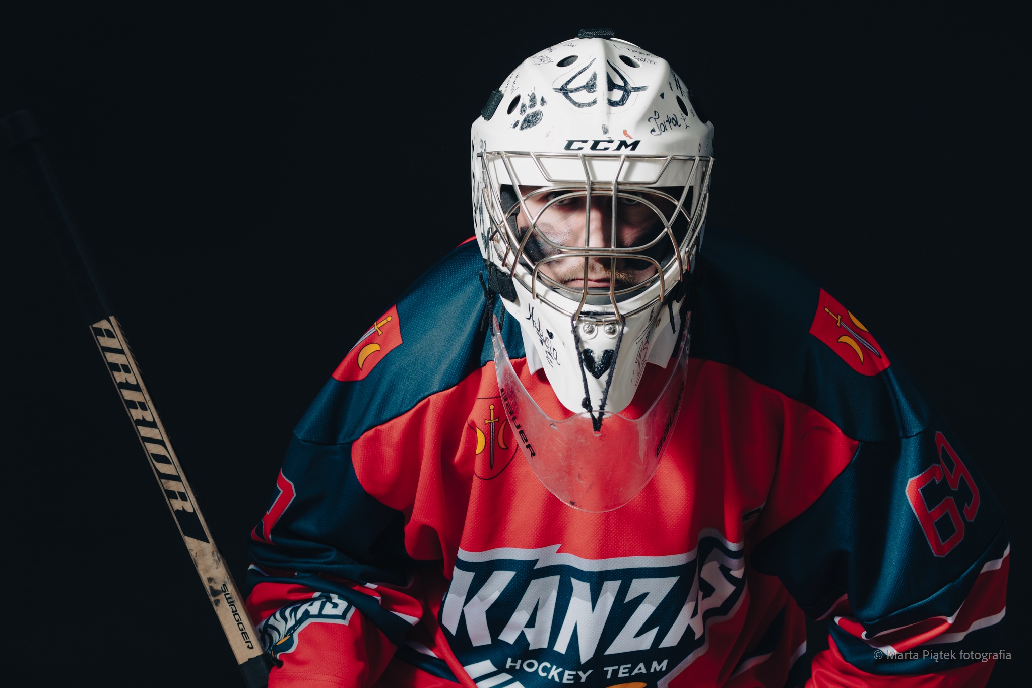 Portret hokeisty w pełnym rynsztunku: kask z rysunkami, czerwono-granatowa koszulka z logo 'KANZA Hockey Team' i numerem 63, trzymającego kij Warrior Swagger na ciemnym tle.