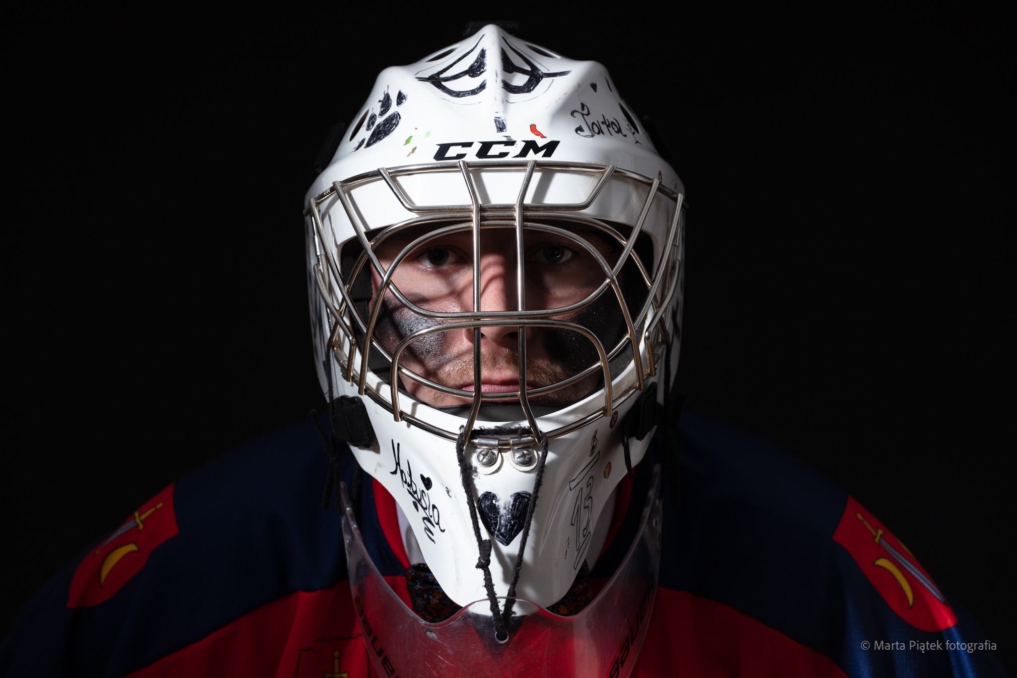 Portret hokejowego bramkarza w masce CCM z autografami i rysunkami, w barwach klubowych na czarnym tle.