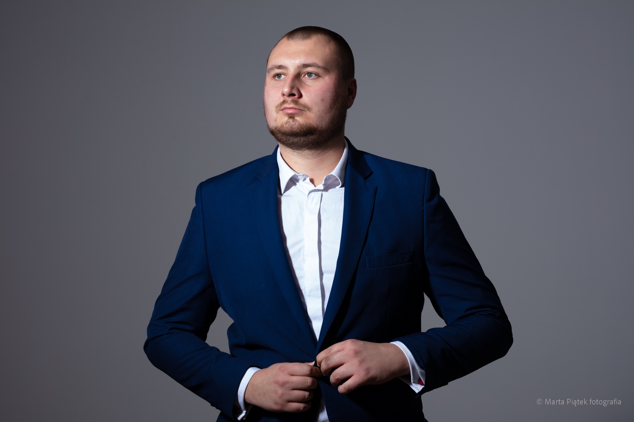 Mężczyzna w granatowym garniturze zapinający guzik, z lekkim zarostem i spojrzeniem w bok na szarym tle.