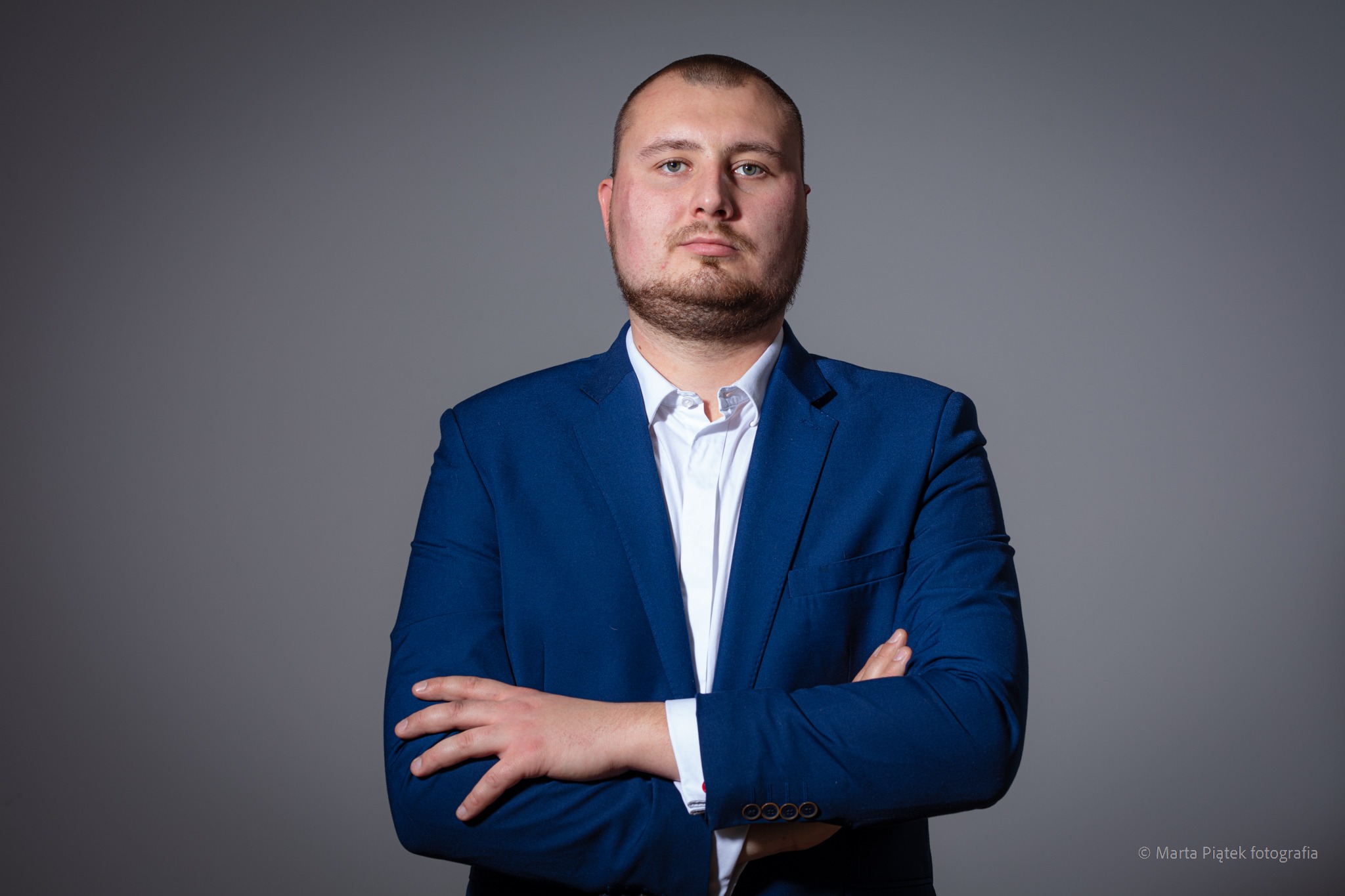 Portret mężczyzny w niebieskim garniturze i białej koszuli, z założonymi rękami na szarym tle, wykonany przez fotografa Martę Piątek.