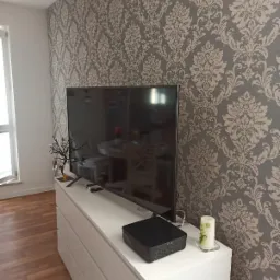 Szara ściana z tapetą w kremowe ornamenty, biała komoda z telewizorem i dekoracjami, minimalistyczny salon.