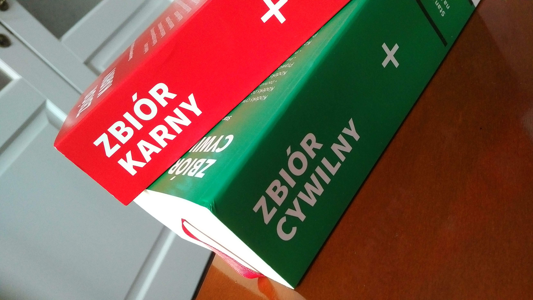 Dwa tomy kodeksów: czerwony 'Zbiór Karny' i zielony 'Zbiór Cywilny', ułożone jeden na drugim, widoczne z bliska na drewnianym stole.