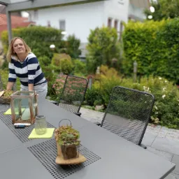 Kobieta w paski opiera się o szary stół na tarasie, na którym stoją dekoracje, w tle zadbany ogród i dom.