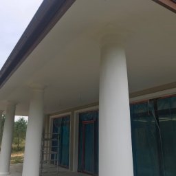 RobotyBudowlane - Białe kolumny i podbitka dachowa po malowaniu w budynku z oknami zabezpieczonymi folią, widoczna drabina.