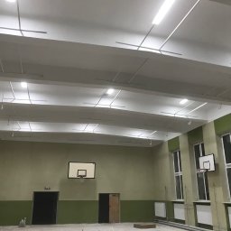 RobotyBudowlane - Sala gimnastyczna po remoncie: biały sufit z widocznymi elementami instalacji, ściany pomalowane na zielono-beżowo, kosze do koszykówki zawieszone na ścianach, nowe oświetlenie.