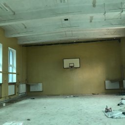 RobotyBudowlane - Wnętrze remontowanej sali gimnastycznej w Białymstoku z widocznymi śladami prac malarskich na ścianach i suficie, oraz sprzętem budowlanym leżącym na podłodze.