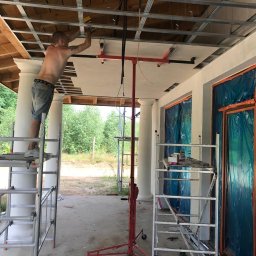 RobotyBudowlane - Mężczyzna na rusztowaniu montuje płytę na metalowym stelażu sufitu z użyciem podnośnika, widoczne kolumny i okna zabezpieczone folią.