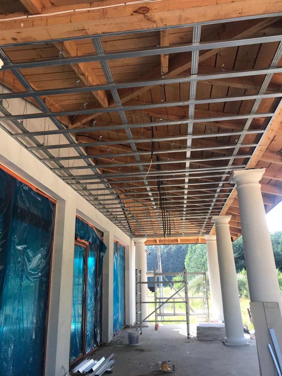 Konstrukcja metalowa podwieszanego sufitu, zamontowana do drewnianej konstrukcji dachu na zewnątrz budynku z kolumnami, okna zabezpieczone folią, widoczna drabina i materiały budowlane.