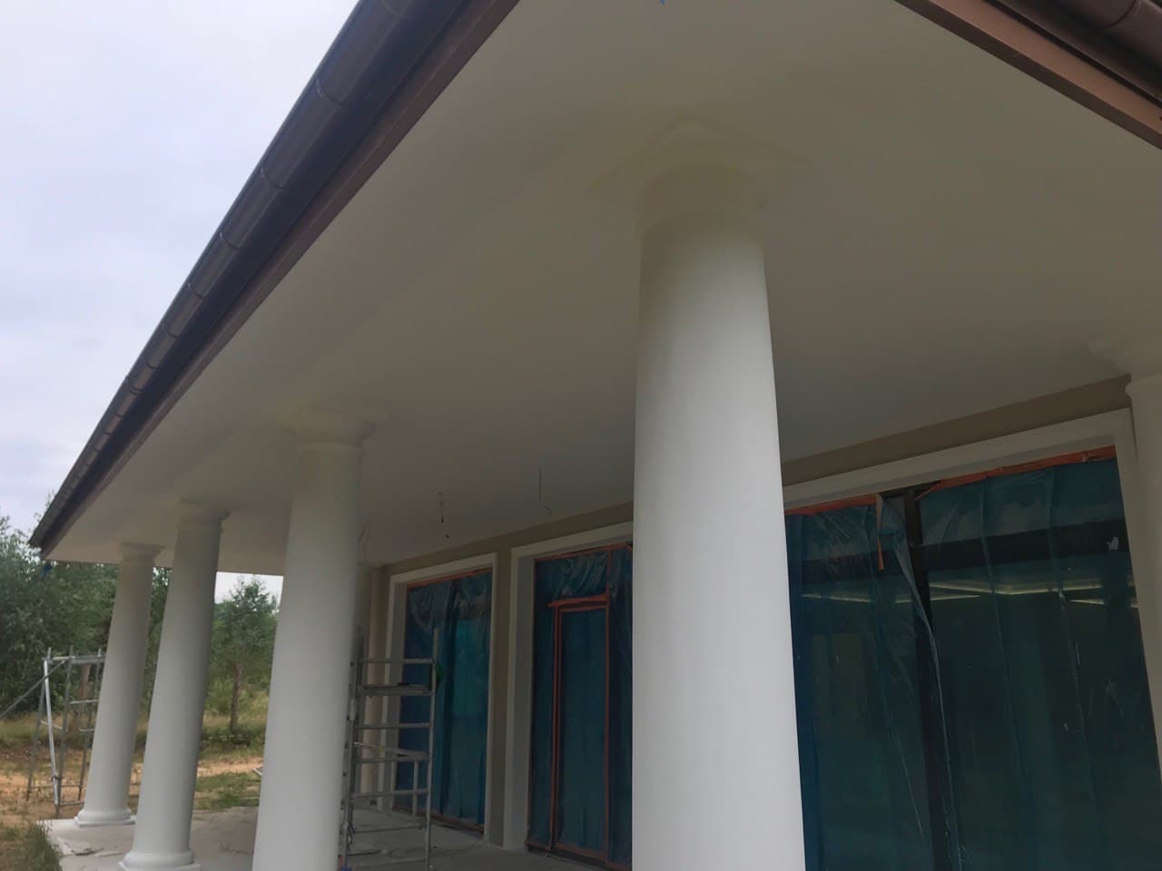 Białe kolumny i podbitka dachowa po malowaniu w budynku z oknami zabezpieczonymi folią, widoczna drabina.