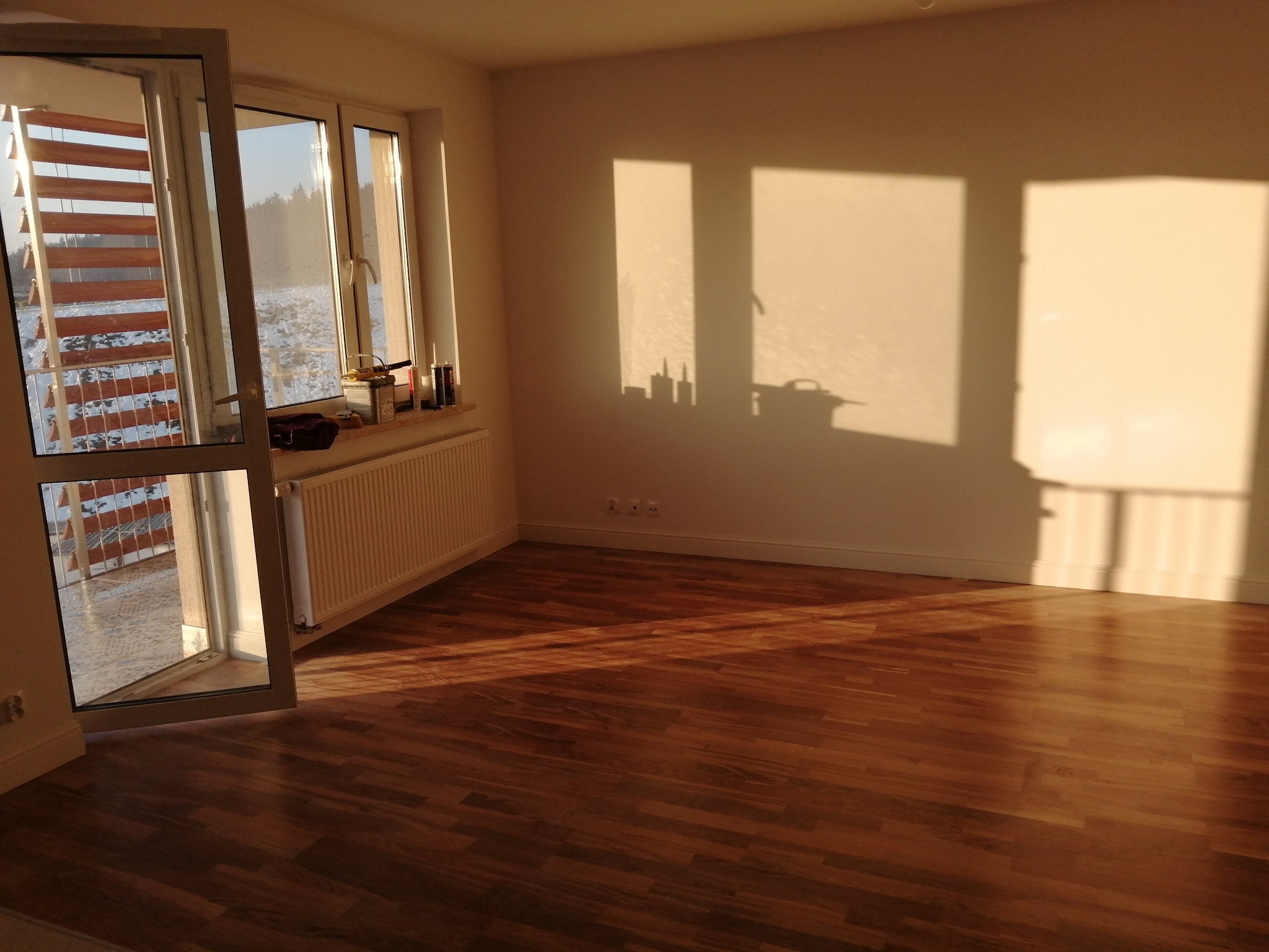 Pusty pokój w Białymstoku po remoncie, z parkietem, oknem balkonowym i promieniami słońca tworzącymi geometryczne cienie na ścianie.