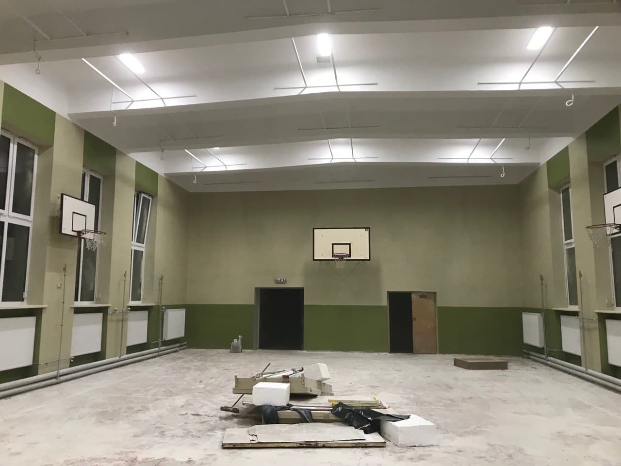 Wnętrze sali gimnastycznej w trakcie remontu: widoczne odświeżone ściany w odcieniach zieleni i beżu, nowe białe sufity z zamontowanym oświetleniem, kosze do koszykówki, okna z grzejnikami,...