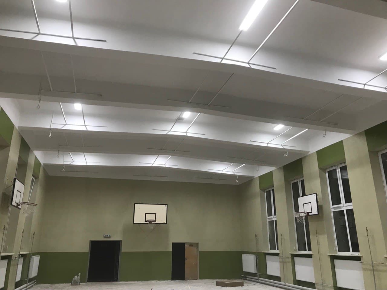 Sala gimnastyczna po remoncie: biały sufit z widocznymi elementami instalacji, ściany pomalowane na zielono-beżowo, kosze do koszykówki zawieszone na ścianach, nowe oświetlenie.