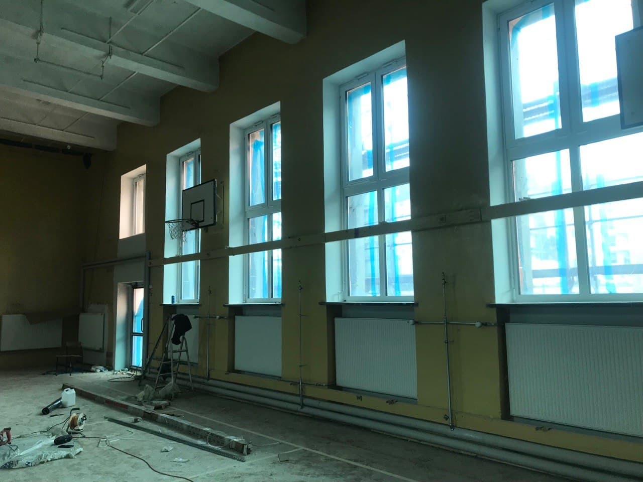 Wnętrze sali gimnastycznej w trakcie remontu: widoczne okna zabezpieczone folią, grzejniki, drabina, narzędzia i ślady prac na podłodze, kosz do koszykówki na ścianie, żółte ściany i biały sufit.