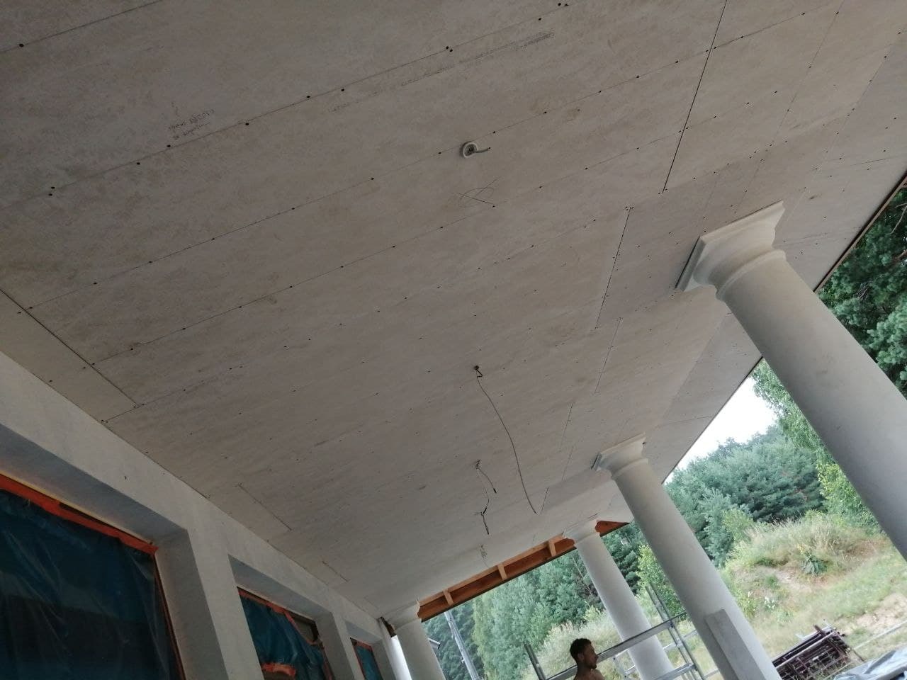 Płyty na suficie werandy z kolumnami w trakcie montażu, widoczne łączenia płyt, wystające kable i osoba na rusztowaniu w tle.