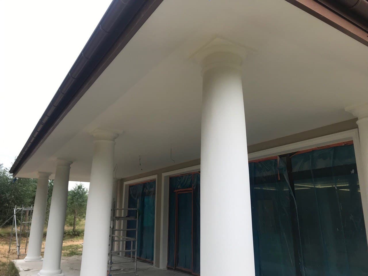 Białe kolumny podtrzymujące poddasze domu jednorodzinnego, zabezpieczone folią okna, drabiny w tle, widok z dołu.