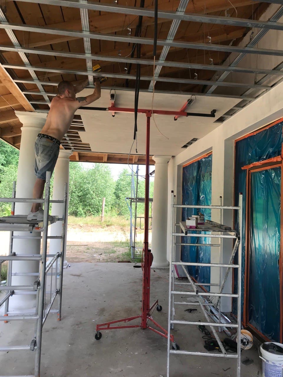 Mężczyzna na rusztowaniu montuje płytę na metalowym stelażu sufitu z użyciem podnośnika, widoczne kolumny i okna zabezpieczone folią.
