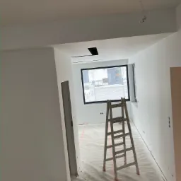 Wnętrze w trakcie prac wykończeniowych: białe ściany, aluminiowa drabina na folii ochronnej, widoczne puszki z farbą i okno z widokiem na otoczenie.