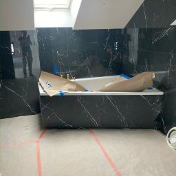 Renovigo.tech - Biała wanna zabezpieczona papierem i niebieską taśmą malarską w łazience z czarnymi marmurowymi płytkami, widoczne okno dachowe i odbicie osoby robiącej zdjęcie.
