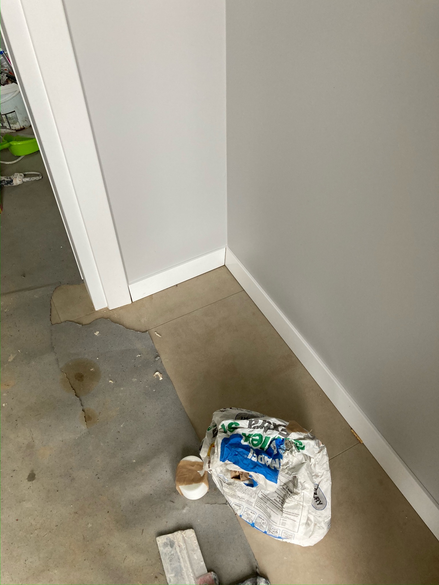 Wnętrze w trakcie prac wykończeniowych: ściana pomalowana na szaro z białymi listwami przypodłogowymi, na podłodze resztki materiałów budowlanych, pęknięta podłoga i worek po zaprawie.
