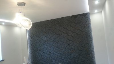 Ściana pokryta dekoracyjną szarą strukturą z geometrycznym wzorem, oświetlona designerską lampą wiszącą i punktowymi światłami w suficie.