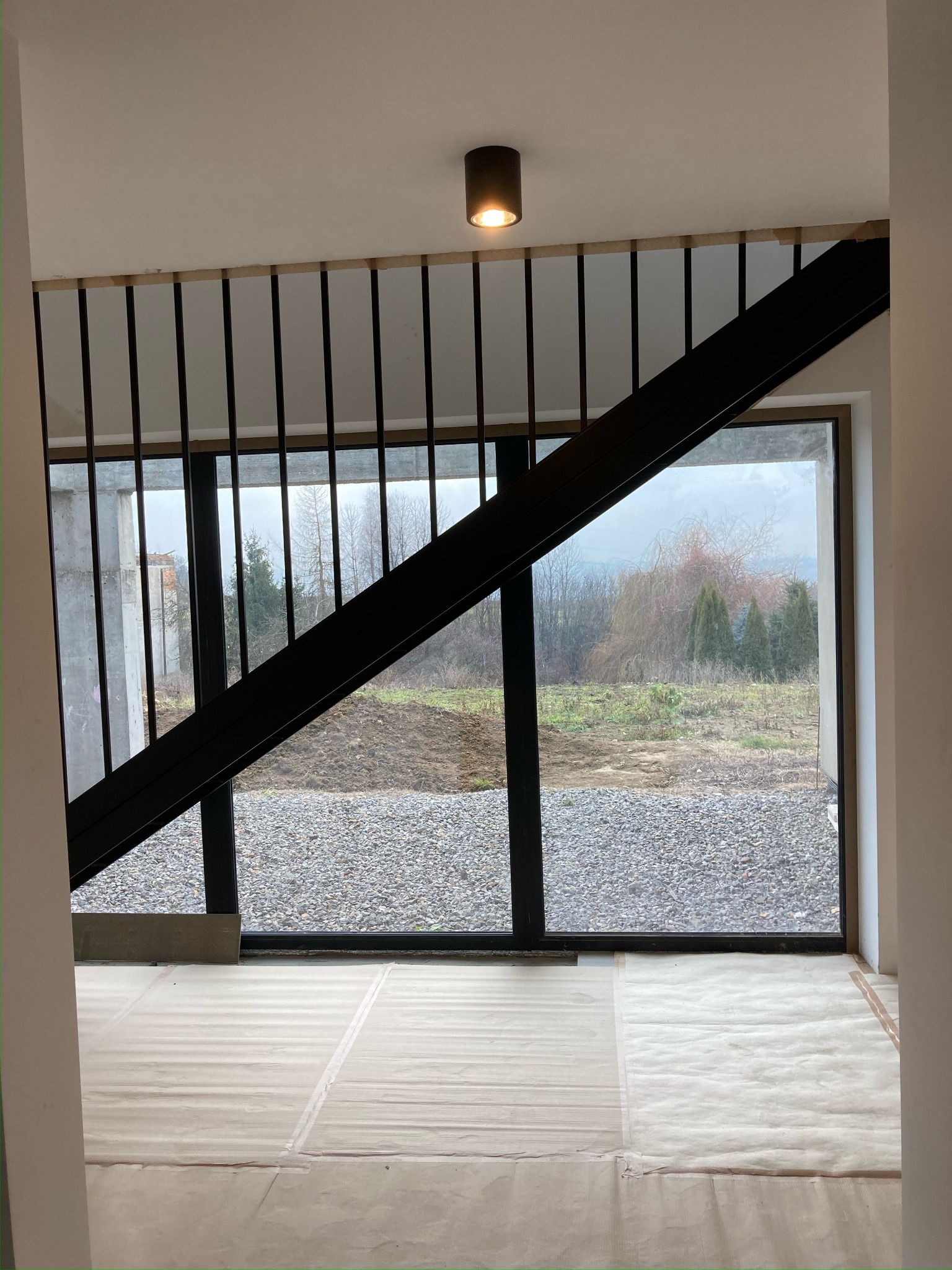 Wnętrze w trakcie wykańczania: widok przez duże okno na zewnątrz, podłoga zabezpieczona folią malarską, surowe ściany i czarna metalowa balustrada schodów.