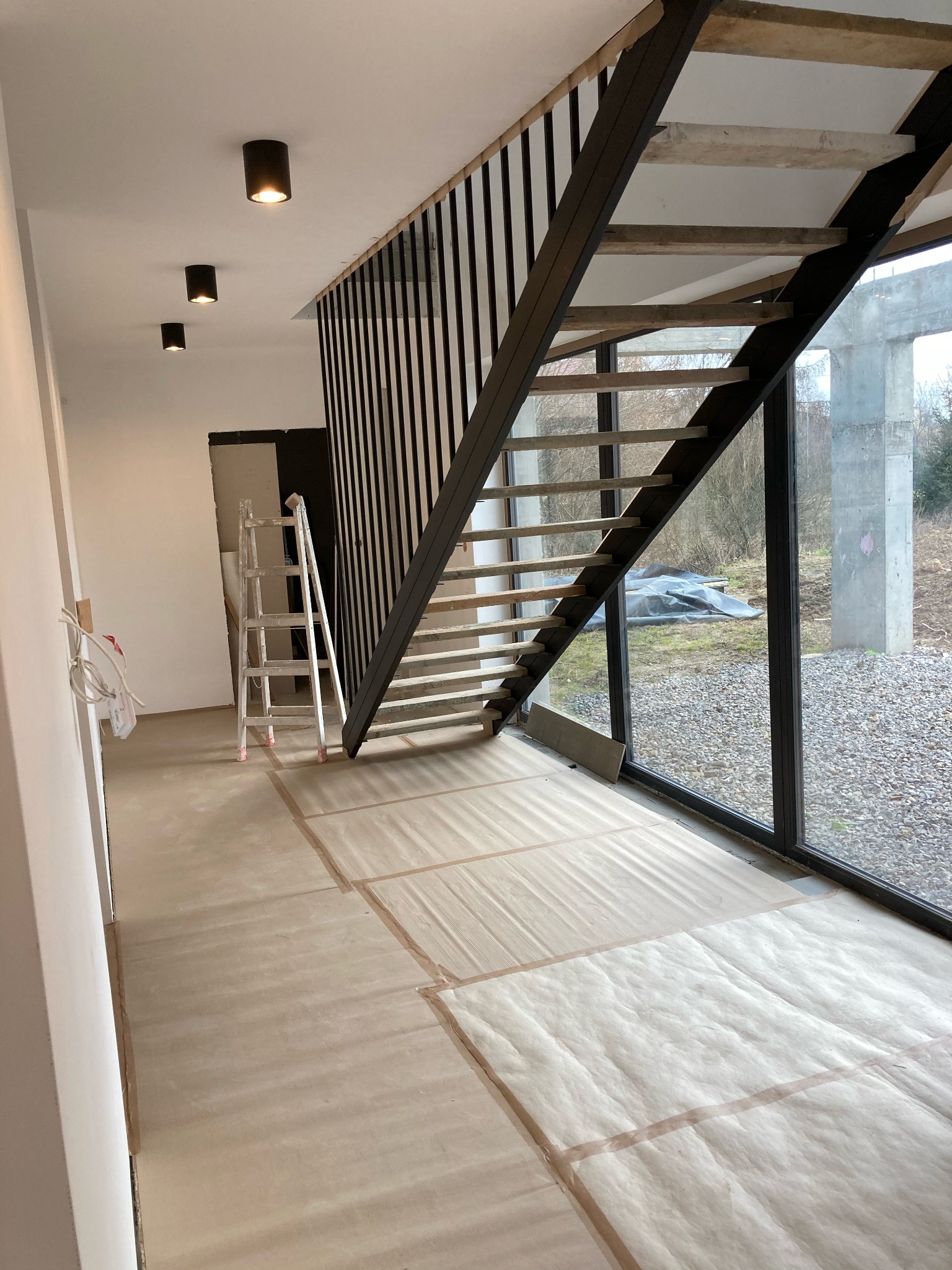 Wnętrze w trakcie prac wykończeniowych: zabezpieczona podłoga, surowe drewniane schody z czarną balustradą, aluminiowa drabina oparta o ścianę, widok na zewnątrz przez duże okno.