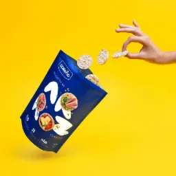 Lekka, niebieska paczka chrupiących wafli ryżowych Costeño w locie na jaskrawym żółtym tle, z dłonią podającą jeden wafel.