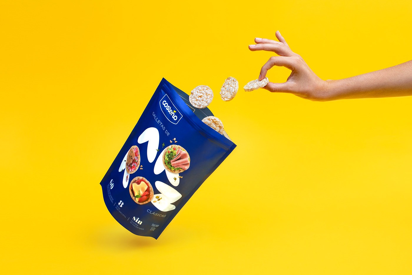Lekka, niebieska paczka chrupiących wafli ryżowych Costeño w locie na jaskrawym żółtym tle, z dłonią podającą jeden wafel.