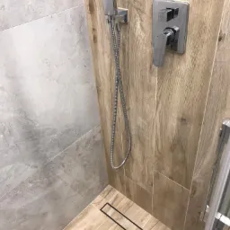 Nowoczesna kabina prysznicowa z odpływem liniowym, wykończona płytkami imitującymi drewno i szarym kamieniem, z chromowaną armaturą.