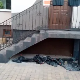 Granitowe schody wejściowe z kutą balustradą, przed remontem, zabezpieczone folią, widoczne donice na ganku.