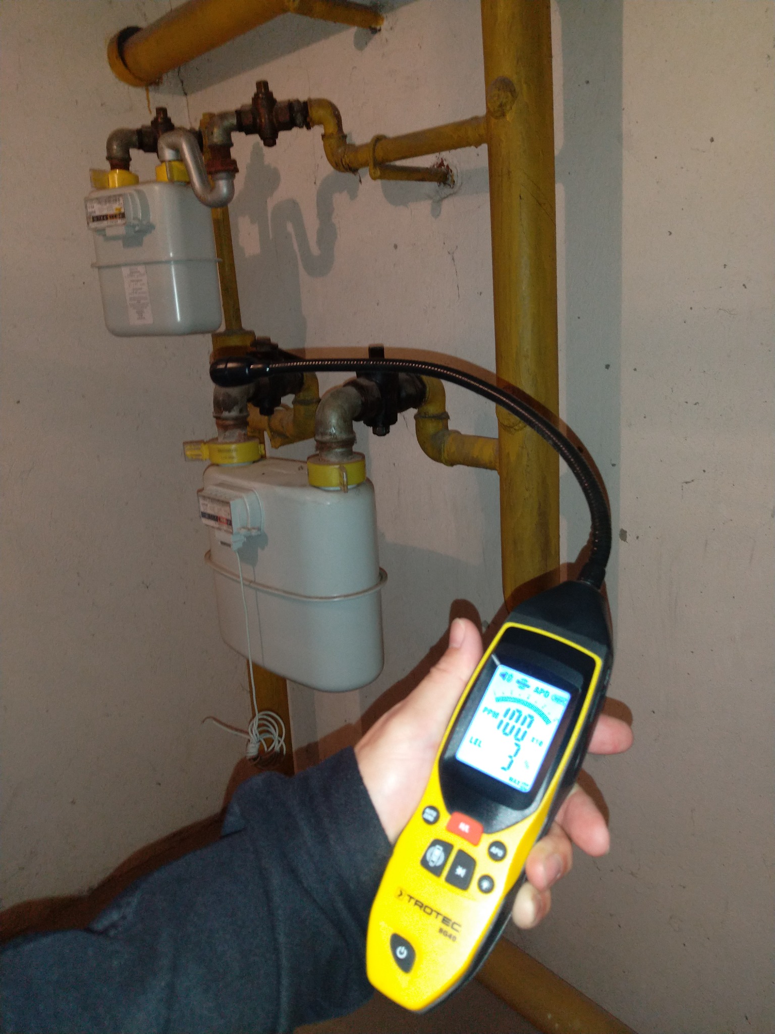 Pomiar szczelności instalacji gazowej: detektor Trotec BG40 w dłoni, rurki gazowe i dwa gazomierze na ścianie w tle. Wyświetlacz detektora pokazuje wartości pomiarowe.