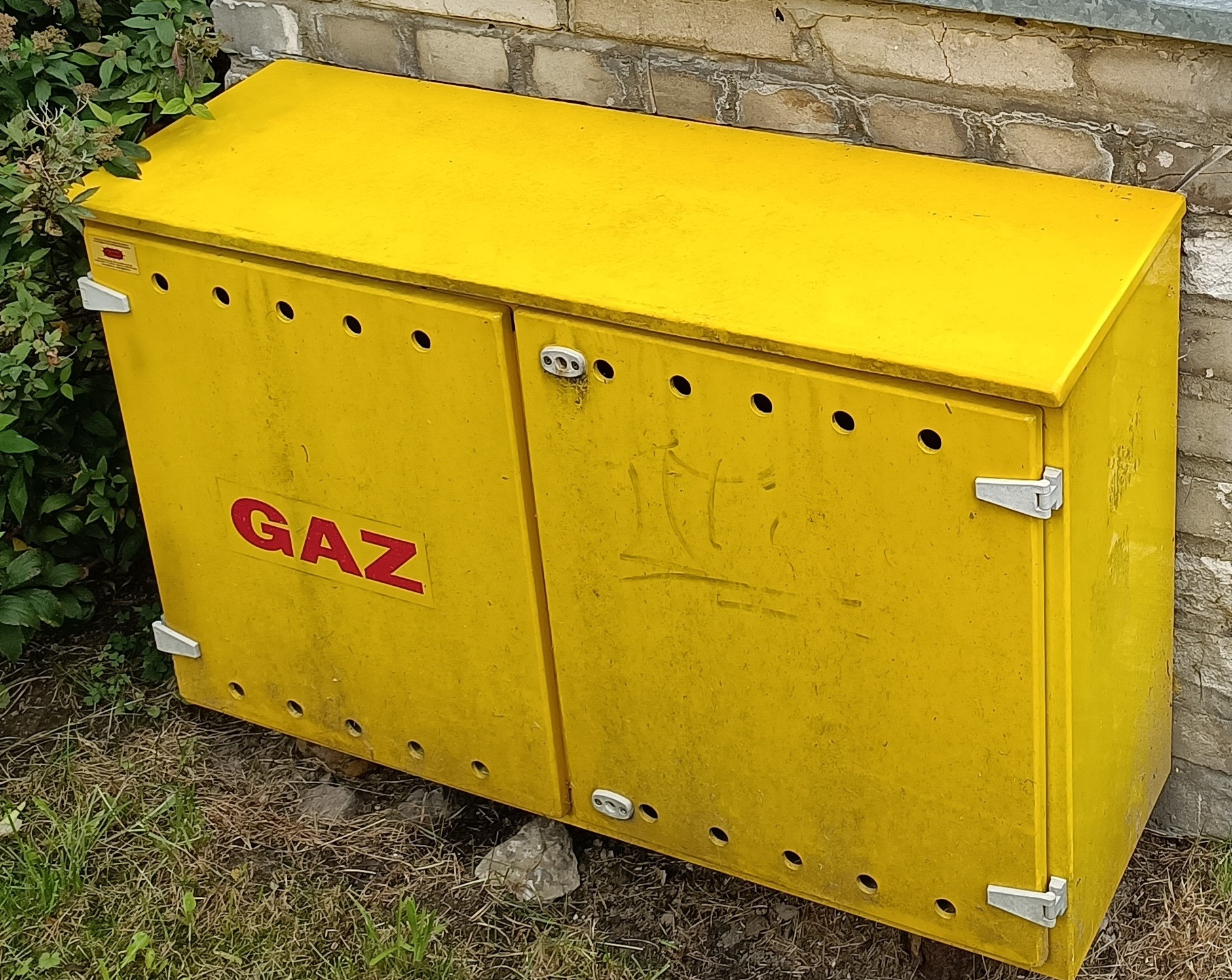 Żółta skrzynka gazowa, z napisem 'GAZ' na drzwiach, umieszczona na zewnątrz budynku przy ceglanej ścianie, otoczona zielenią.