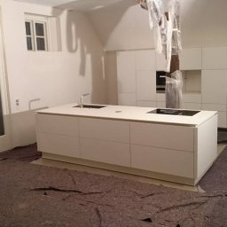 Go Kitchens Tomasz Wojciuk - Montaż białej, minimalistycznej kuchni z wyspą kuchenną i zabudową po sufit, widoczne zabezpieczenia na filarze, podłoga zabezpieczona kartonem, duże przeszklone drzwi balkonowe.