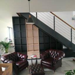 Go Kitchens Tomasz Wojciuk - Pokój dzienny z dwoma bordowymi skórzanymi fotelami typu chesterfield, pufem, regałem z czarnymi i surowymi drewnianymi półkami pod schodami z metalową balustradą, lampą wiszącą i roślinami...