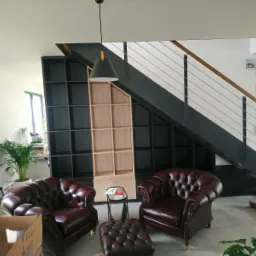 Pokój dzienny z dwoma bordowymi skórzanymi fotelami typu chesterfield, pufem, regałem z czarnymi i surowymi drewnianymi półkami pod schodami z metalową balustradą, lampą wiszącą i roślinami...