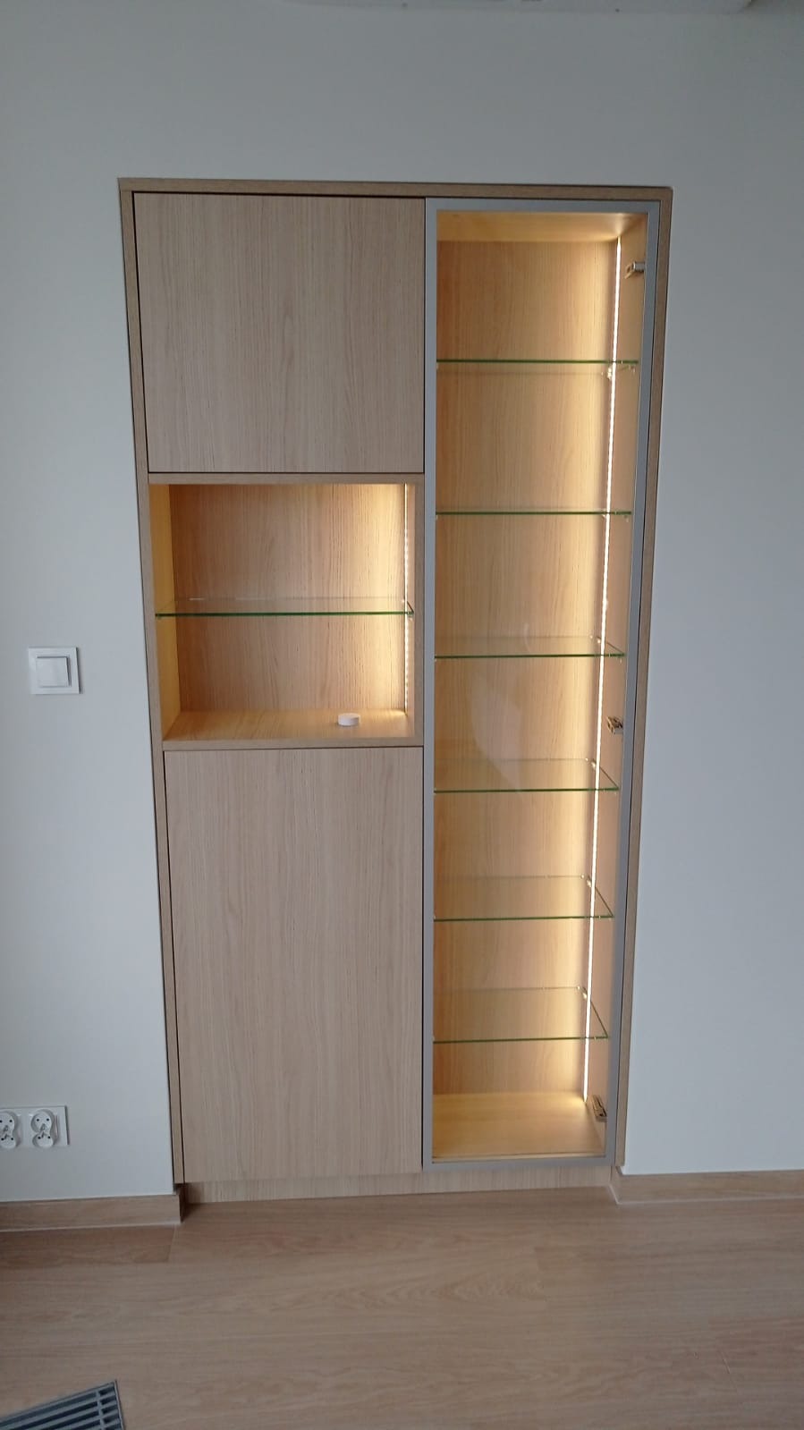 Jasna, drewniana szafa z przeszkloną witryną i wewnętrznym oświetleniem LED, z półkami z przezroczystego szkła, zamontowana na ścianie z widocznymi gniazdkami elektrycznymi.