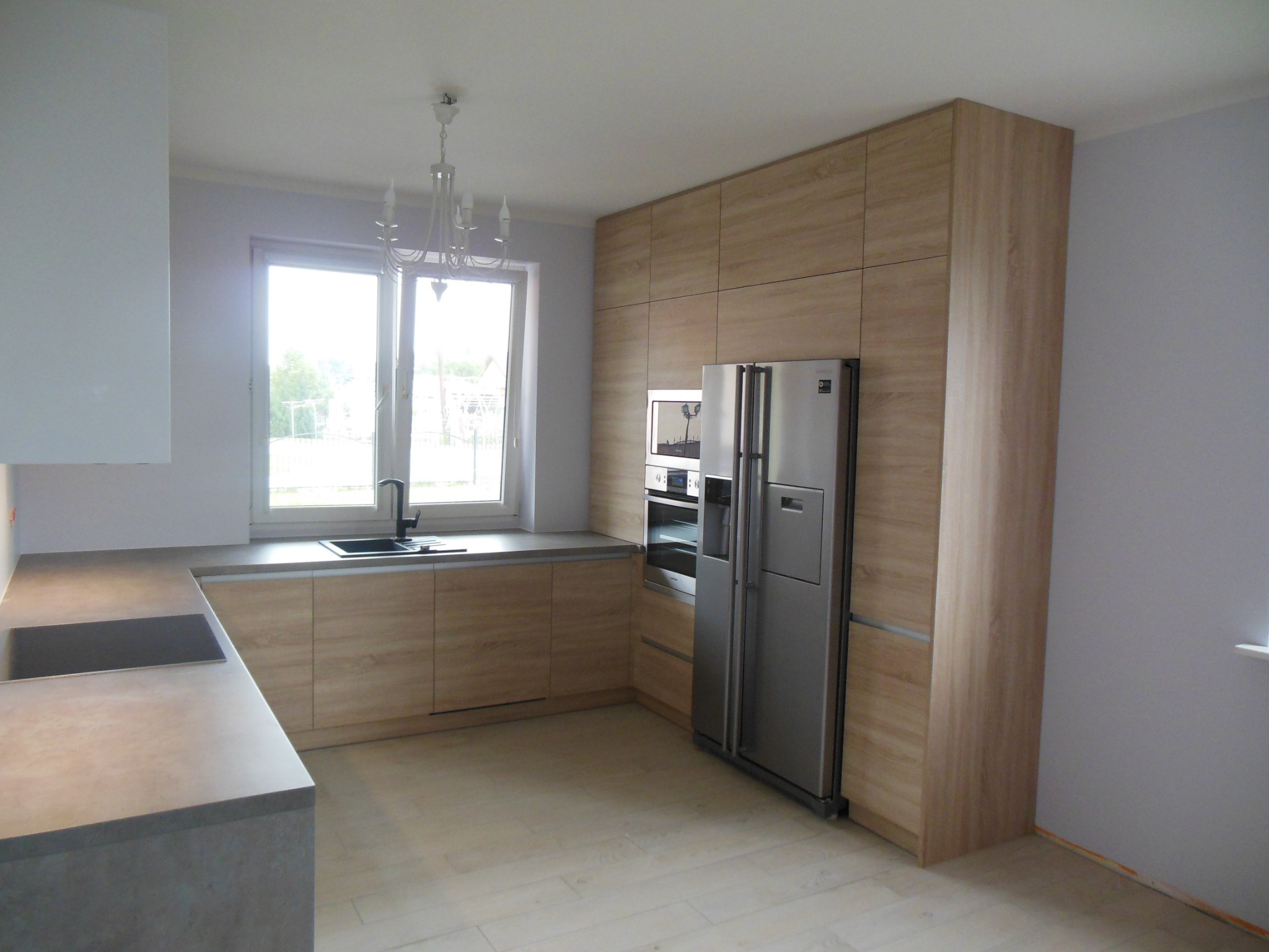 Jasna kuchnia z drewnianymi frontami, szarym blatem i srebrną lodówką side-by-side, oświetlona światłem dziennym z okna z żyrandolem.