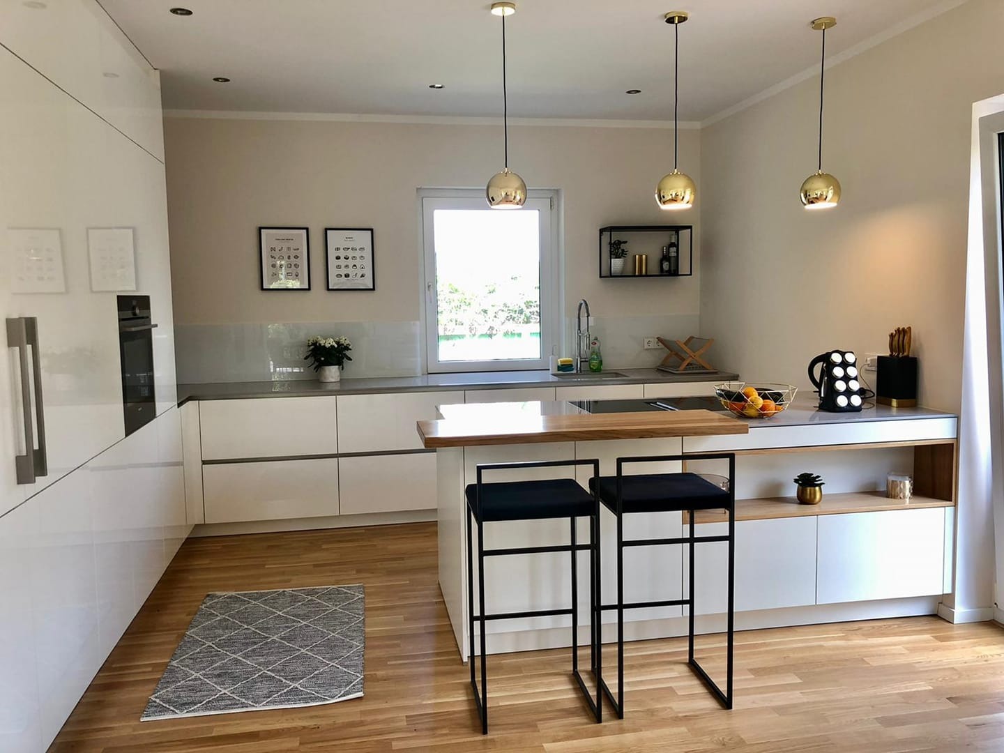 Nowoczesna kuchnia z białymi, lakierowanymi frontami, drewnianym blatem wyspowym i czarnymi hokerami na tle parkietu; złote lampy wiszące nad wyspą.