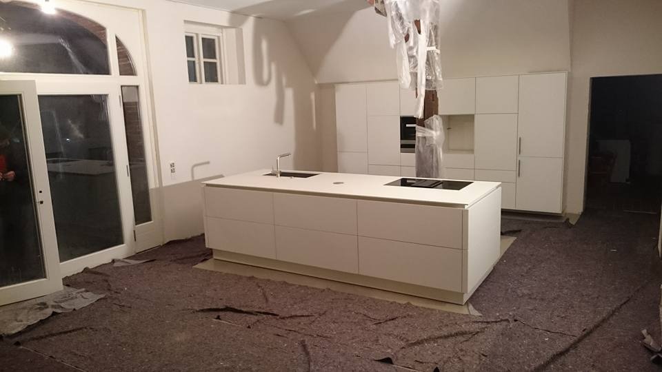 Montaż białej, minimalistycznej kuchni z wyspą kuchenną i zabudową po sufit, widoczne zabezpieczenia na filarze, podłoga zabezpieczona kartonem, duże przeszklone drzwi balkonowe.