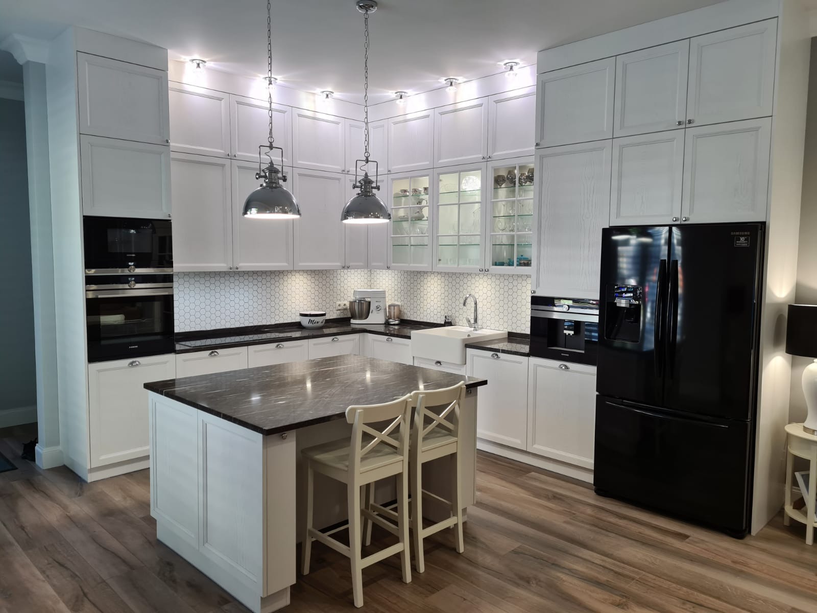 Jasna kuchnia z białymi szafkami, czarnym blatem i wyspą kuchenną z dwoma krzesłami, oświetlona wiszącymi lampami w stylu industrialnym.