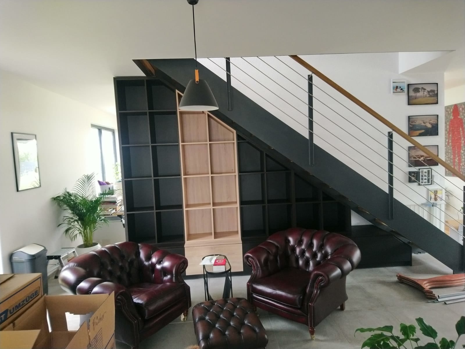 Pokój dzienny z dwoma bordowymi skórzanymi fotelami typu chesterfield, pufem, regałem z czarnymi i surowymi drewnianymi półkami pod schodami z metalową balustradą, lampą wiszącą i roślinami...