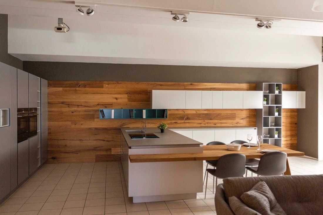 Nowoczesna kuchnia z szarymi szafkami, drewnianą ścianą i wyspą z blatem łączącym się z drewnianym stołem jadalnym. Na stole nakrycia i kieliszki do wina.