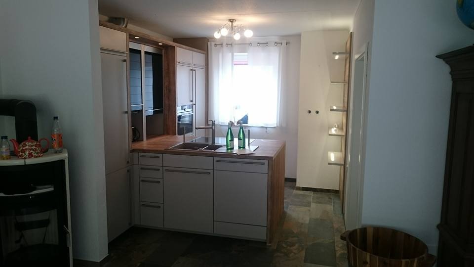 Jasna kuchnia z wyspą i drewnianym blatem, widoczne butelki wody mineralnej na blacie, w tle okno z białą zasłoną i lampa sufitowa.