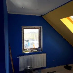 Pokój na poddaszu w trakcie malowania: ściany pomalowane na intensywny niebieski, fragment sufitu przy oknie dachowym w kolorze żółtym, widoczne narzędzia malarskie przy oknie, grzejnik centralnego...