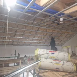 Poddasze w trakcie ocieplania: widoczne rolki wełny mineralnej Ursa Gold 35, metalowy stelaż sufitu i niebieskie rury wentylacyjne. Prace wykończeniowe.