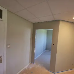 Świeżo pomalowane wnętrze biura w odcieniach szarości, z zabezpieczonymi listwami przypodłogowymi niebieską taśmą malarską i widocznym fragmentem sufitu kasetonowego.