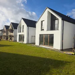 Nowoczesne domy jednorodzinne z białymi elewacjami i ciemnymi dachami, jeden dom w trakcie prac elewacyjnych, widoczne rusztowania i trawnik.