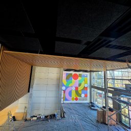 MICHAŁ WITTBRODT - Wnętrze w trakcie wykończenia: drewniane lamele na ścianie, kolorowa grafika na ścianie, pracownik na rusztowaniu montuje panele akustyczne na suficie.