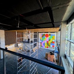 MICHAŁ WITTBRODT - Prace wykończeniowe w nowoczesnym wnętrzu: malowanie sufitu z rusztowania, widoczna kolorowa grafika na ścianie i duże okna z widokiem na zewnątrz.