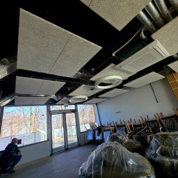 MICHAŁ WITTBRODT - Montaż paneli akustycznych na suficie w nowym biurze. Widoczne instalacje, oświetlenie i pracownik przy oknie. Krzesła w folii czekają na ustawienie.